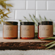 Load image into Gallery viewer, Herbal Bath Salts in amber jars. There is a relaxing bath salt, reviving bath salt, and seduction bath salt. They are placed on a block of wood in front of subway tiles and a palm tree.
