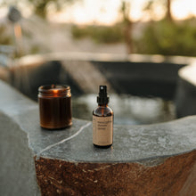 Load image into Gallery viewer, Essential oil based eucalyptus shower spray in a 2 oz amber glass bottle in an outdoor hot springs bath next to a burning candle. The water is running behind the product.
