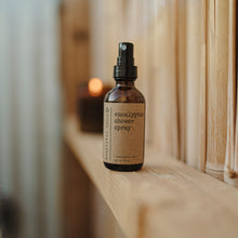 Load image into Gallery viewer, Essential oil based eucalyptus shower spray in a 2 oz amber glass bottle in an outdoor wooden shower next to a burning candle.
