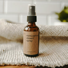Load image into Gallery viewer, Essential oil based eucalyptus shower spray in a 2 oz amber glass bottle on top of a white blanket in front of subway tiles and a plant.
