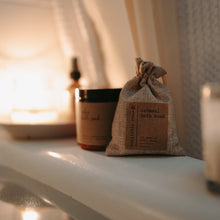 Load image into Gallery viewer, A lifestyle shot of an organic herbal oatmeal bath soak in a burlap bag, a relaxing epsom salt bath with lavender and chamomile, and a shower spray all in a bath tub full of water. There is also a lit candle.
