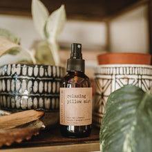 Load image into Gallery viewer, Relaxing pillow mist in a 4 oz amber glass bottle on a shelf next to decorative plants and a palo santo burning.
