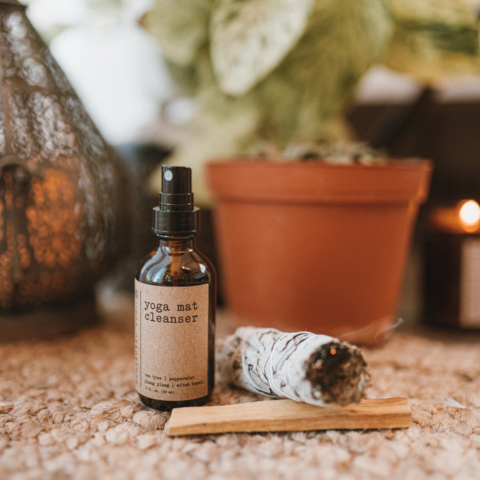 Yoga mat cleanser with rose quartz gemstones in a 2 oz glass amber bottle sitting on the floor with a sage bundle burning and palo santo stick. The products are in front of a plant and a lantern with a burning candle.