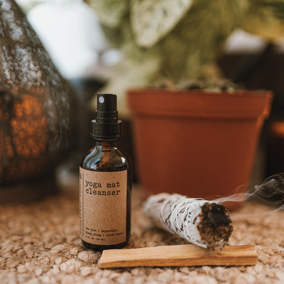Yoga mat cleanser with rose quartz gemstones on the floor with a burning sage bundle & palo santo stick. In front of a plant, lantern & burning candle.