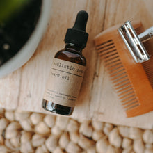 Load image into Gallery viewer, Beard oil in an amber bottle with glass dropper next to a wooden comb and silver straight razor. 
