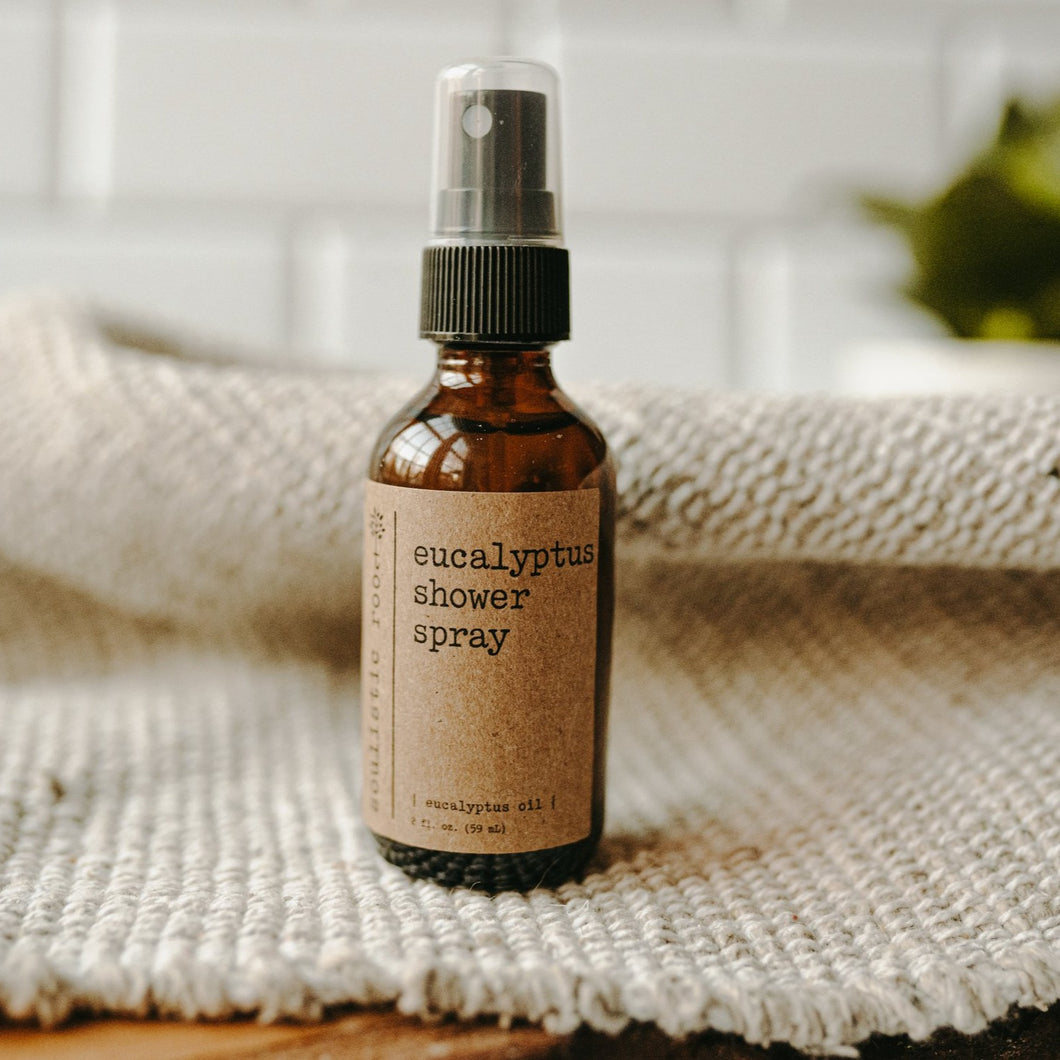 Essential oil based eucalyptus shower spray in a 2 oz amber glass bottle on top of a white blanket in front of subway tiles and a plant.
