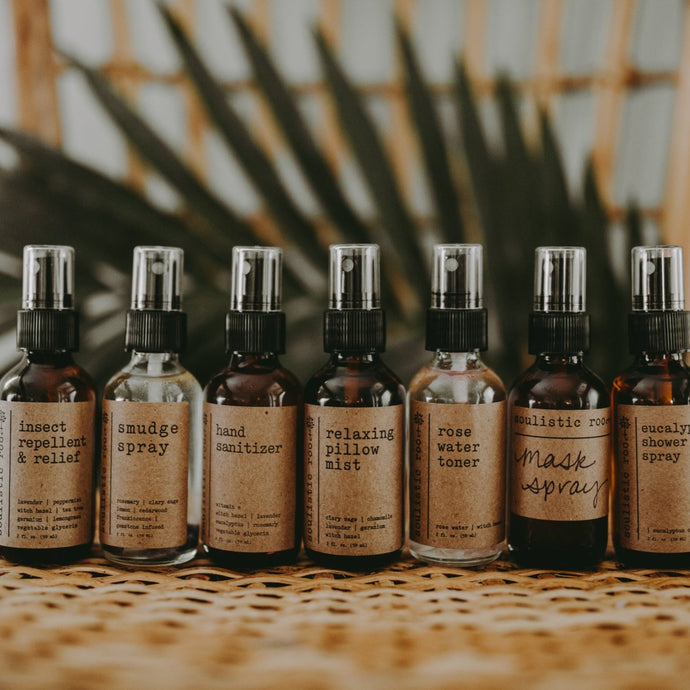 A group shot of products on a wood bench in front of a plant. The products included are- bug spray, smudge spray, hand sanitizer, relaxing pillow mist, rose water toner, essential oil mask spray, and eucalyptus shower spray.