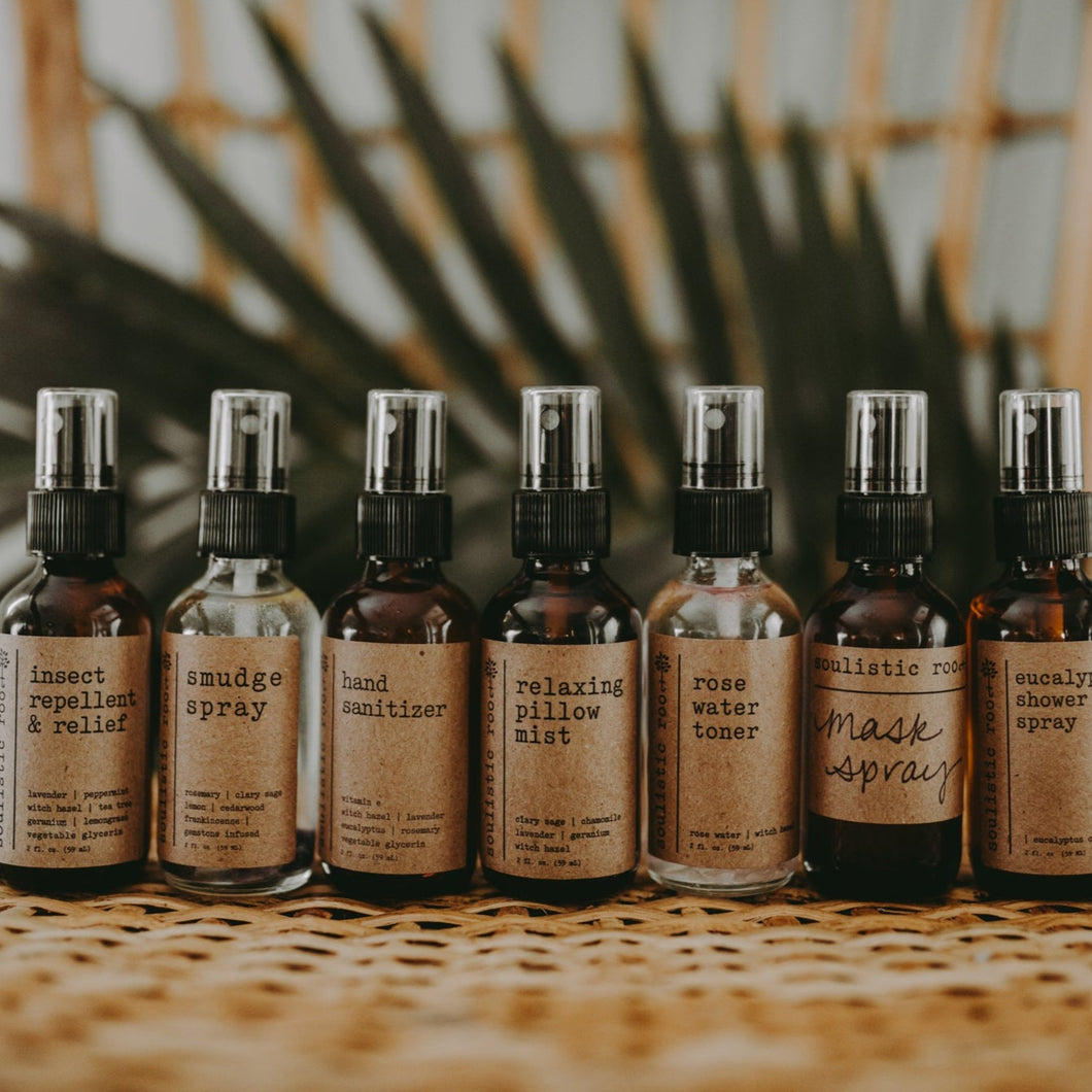 A group shot of products on a wood bench in front of a plant. The products included are- bug spray, smudge spray, hand sanitizer, relaxing pillow mist, rose water toner, essential oil mask spray, and eucalyptus shower spray.