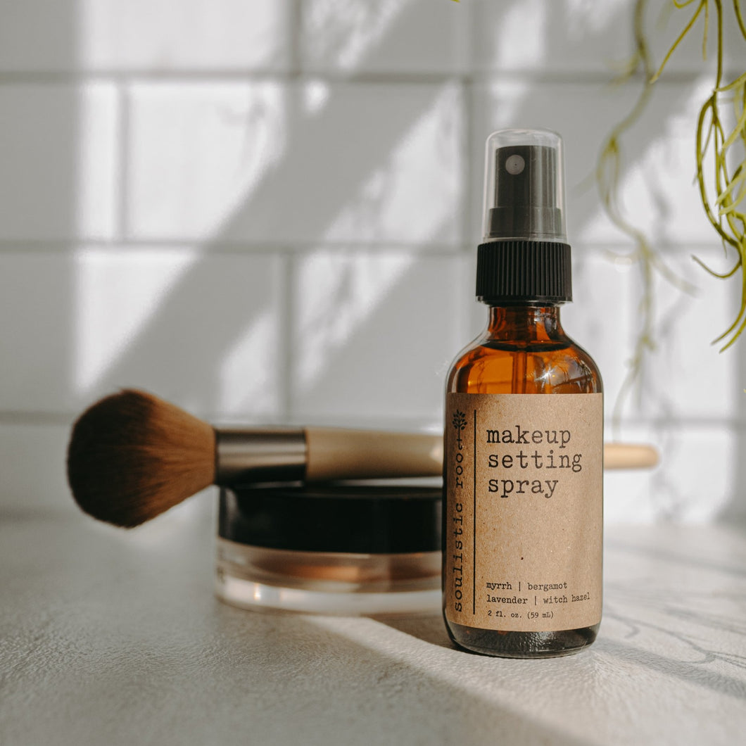 Makeup setting spray in a 2 oz amber glass bottle in front of loose powder and a makeup brush and subway tiles.