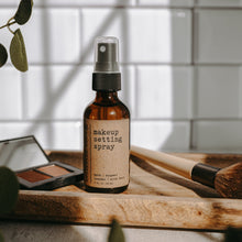 Load image into Gallery viewer, A makeup setting spray in a 2 oz amber glass bottle on a piece of wood with a makeup brush and some eyeshadow in front of subway tile.
