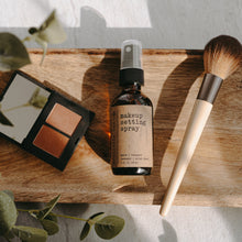Load image into Gallery viewer, A makeup setting spray in a 2 oz amber glass bottle on a piece of wood with a makeup brush and some eyeshadow.
