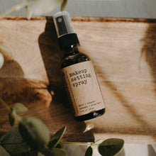 Load image into Gallery viewer, Makeup setting spray in a 2 oz amber glass bottle in front of a piece of wood next to a eucalytpus plant.
