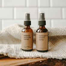 Load image into Gallery viewer, Makeup setting spray and a eucalyptus shower spray on a piece of wood with a white blanket in front of subway tiles. Both products are in a 2 oz amber glass spray bottle.
