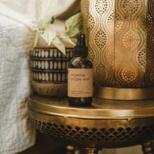 Load image into Gallery viewer, Relaxing essential oil pillow mist in a 4 oz amber glass bottle on a side table with plants and candles next to a bed.
