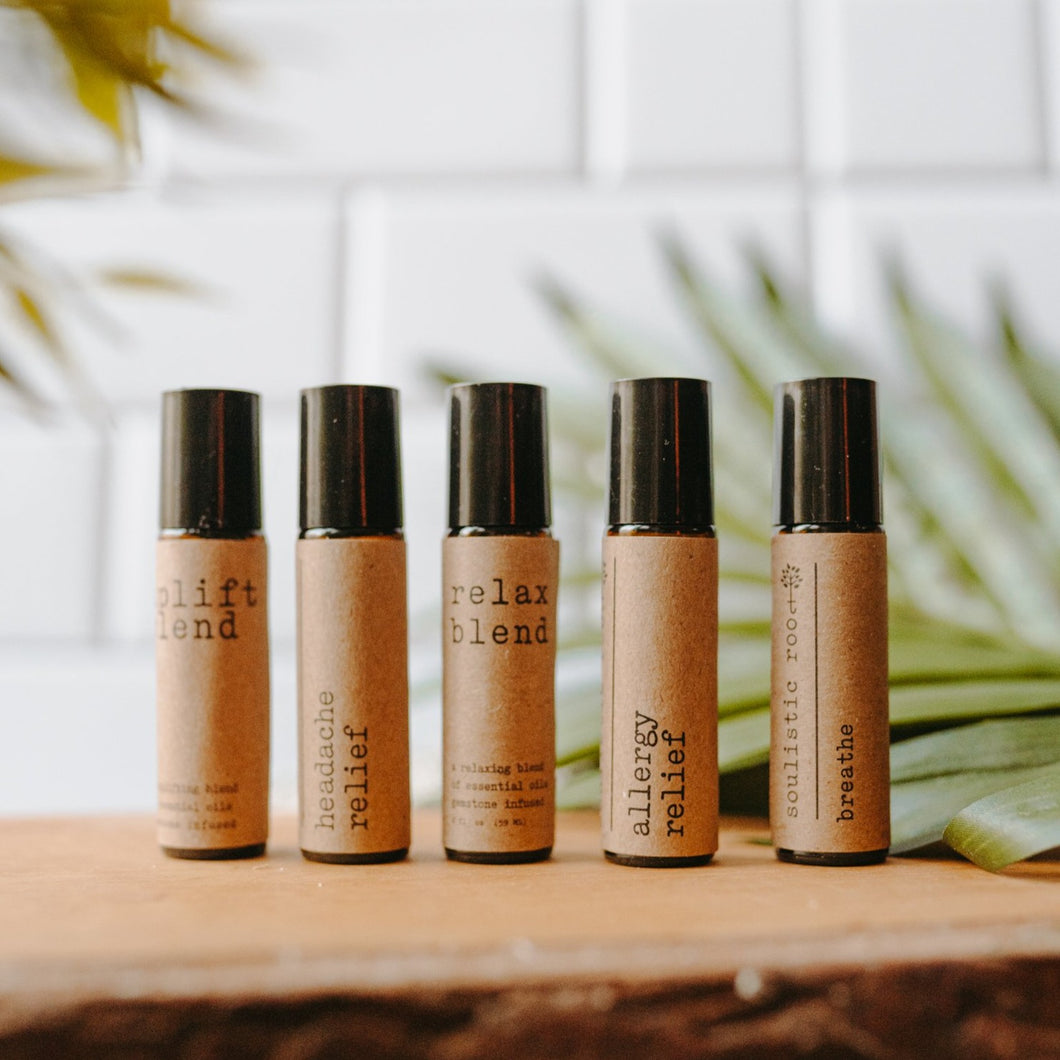 Essential oil rollers. There are five different blends -uplift which has rose quartz gemstones, headache relief, relax blend which has amethyst gemstones, allergy relief and breathe. They are in amber roller bottles on a wood block in front of a palm leaf.