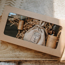 Load image into Gallery viewer, A travel set full of sample sizes already wrapped in a kraft box. The set includes a lavender &amp; chamomile epsom salt bath, essential oil headache relief roller, organic herbal oatmeal bath soak &amp; a relaxing pillow spray. The set is on a piece of wood on a white blanket.
