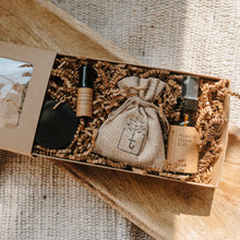 Load image into Gallery viewer, A travel set full of sample sizes already wrapped in a kraft box. The set includes a lavender &amp; chamomile epsom salt bath, essential oil headache relief roller, organic herbal oatmeal bath soak &amp; a relaxing pillow spray. The set is on a piece of wood and blanket in the background.
