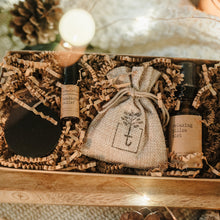 Load image into Gallery viewer, A travel set full of sample sizes already wrapped in a kraft box. The set includes a lavender &amp; chamomile epsom salt bath, essential oil headache relief roller, organic herbal oatmeal bath soak &amp; a relaxing pillow spray. The set is on a piece of wood and blanket with pine cones and lights in the background.
