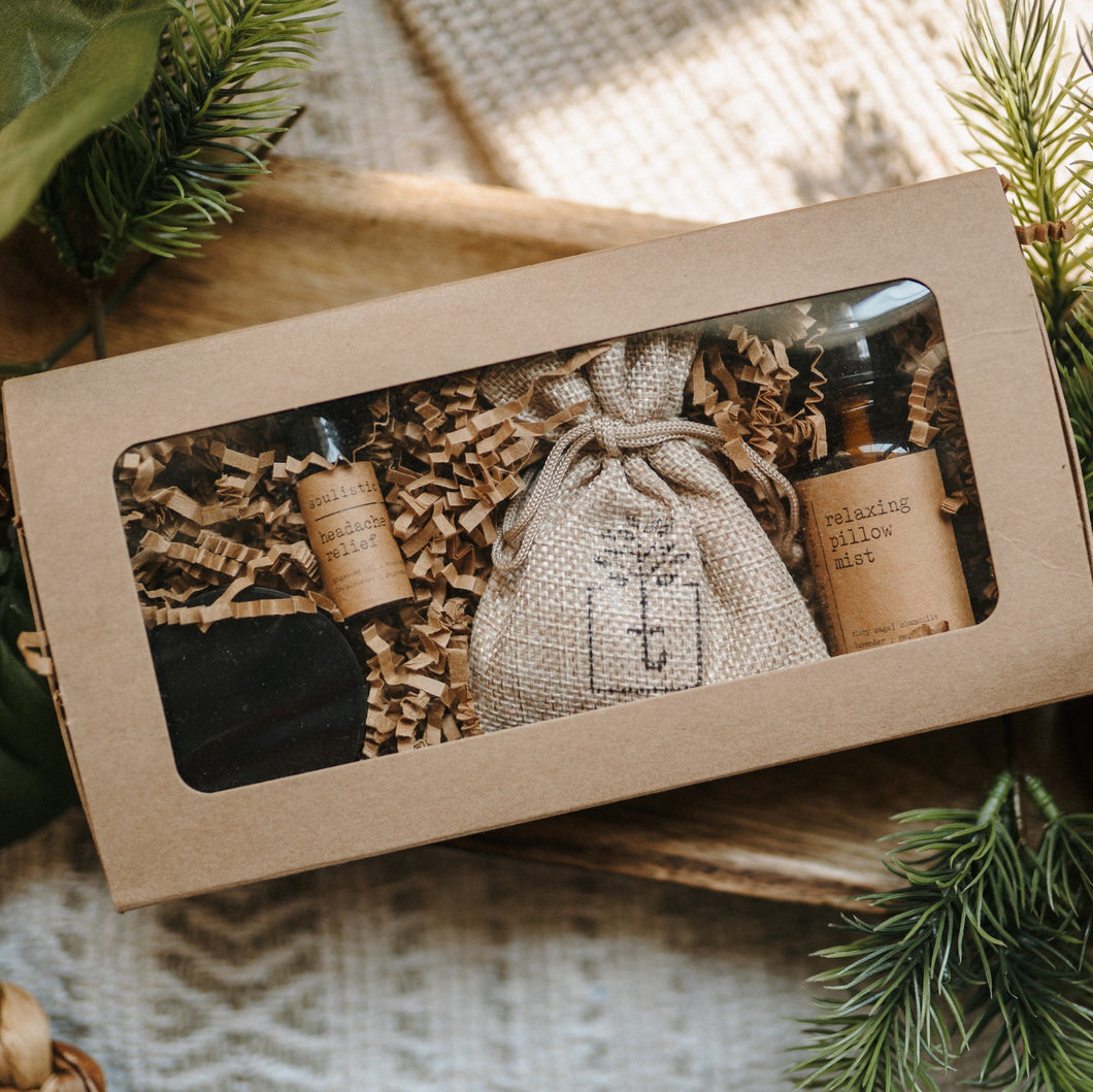 A travel set full of sample sizes already wrapped in a kraft box. The set includes a lavender & chamomile epsom salt bath, essential oil headache relief roller, organic herbal oatmeal bath soak & a relaxing pillow spray. The set is on a piece of wood and blanket with pine in the background.