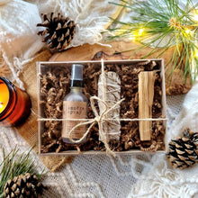 Load image into Gallery viewer, Amethyst infused smudge spray made with essential oils in a gift set with a sage bundle and palo santo stick. Wrapped in a kraft box with krinkle paper and a clear lid with ribbon. The set is on a piece of wood with pinecones on a blanket with lit candles and a lit up tree.
