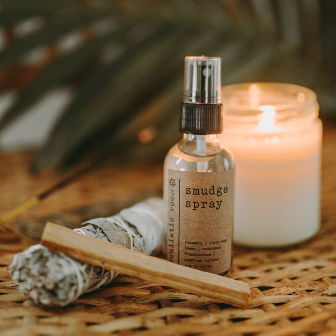 Amethyst Infused Essential Oil Smudge Spray on a wicker table with a sage and palo santo burning. There is also a white candle and palm leaf. 