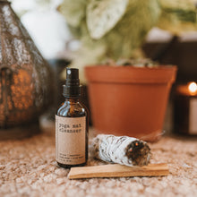 Load image into Gallery viewer, Yoga mat cleanser with rose quartz gemstones in a 2 oz glass amber bottle sitting on the floor with a sage bundle burning and palo santo stick. The products are in front of a plant and a lantern with a burning candle.
